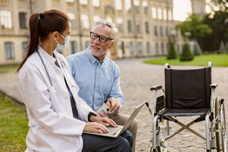 Cheerful mature recovering patient with wheelchair having conversation with young female doctor working on a laptop in the park near clinicの写真素材