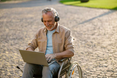 Happy mature man, recovering patient in wheelchair wearing headphones working on a laptop outdoors in the park near clinicの写真素材