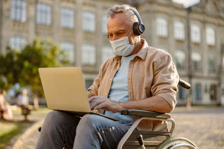 Focused mature man, recovering patient in wheelchair wearing protective mask and headphones working on a laptop outdoors in the park near clinicの写真素材
