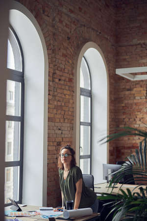 Interior designer or architect in casual wear with messy hairdo looking at camera while working on a blueprint for new project, standing in her office on a daytimeの写真素材