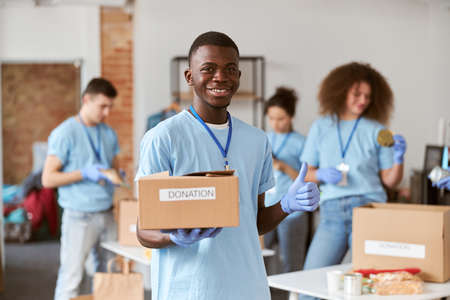 Portrait of african american young male volunteer in blue uniform, protective gloves smiling, showing thumbs up and holding cardboard box for Donationの写真素材