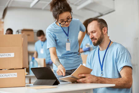 Man and woman working together on donation project indoors. Team of diverse volunteers in blue uniform making notes, calculating, sorting and packing items in cardboard boxesの写真素材