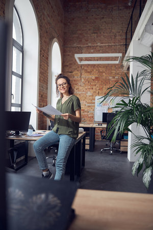 Joyful woman, interior designer or architect wearing glasses smiling at camera while working on a blueprint for new project, leaning on the table in officeの写真素材