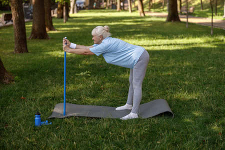 Smiling mature lady in tracksuit does forward bend leaning onto bar in parkの写真素材