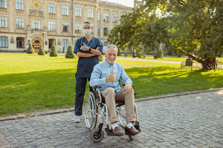 Joyful senior man, recovering patient in a wheelchair smiling at camera, showing thumbs up together with a nurse wearing face shield and mask on a summer day outdoorsの写真素材