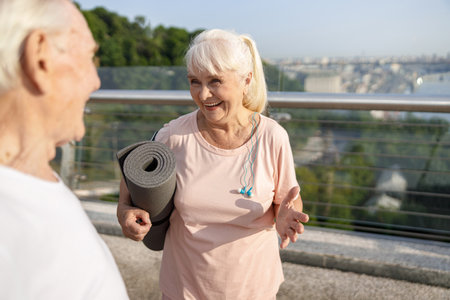 Senior lady with mat and earphones talks to man on footbridgeの写真素材