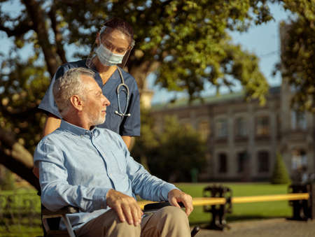 Caring young nurse wearing face shield and mask talking to mature man, pushing patient in wheelchairの写真素材