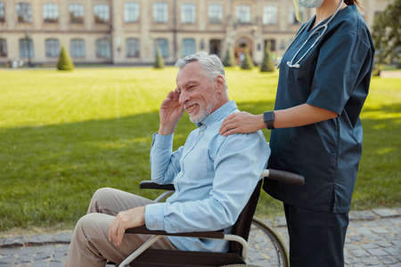 Happy aged man, recovering patient in a wheelchair on a walk with a nurse wearing face shield and mask on a summer day outdoorsの写真素材