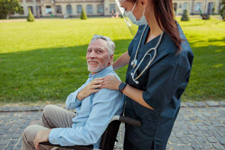Caring nurse wearing face shield and mask on a walk with aged man, recovering patient in a wheelchair on a summer day outdoorsの写真素材