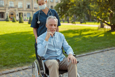 Senior man, recovering patient in a wheelchair looking at camera while on a walk with a nurse wearing face shield and mask on a summer day outdoorsの写真素材