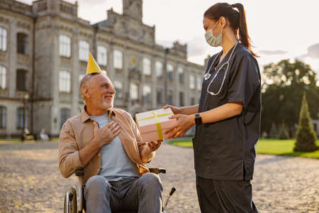 Pleased mature man, recovering patient in wheelchair wearing birthday hat receiving gift and congratulations from young nurseの写真素材