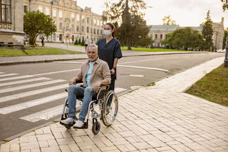 Young nurse in protective face mask assisting senior handicapped man in wheelchair during a walk in...