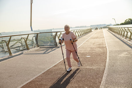 Joyful mature woman in pink tracksuit practices Nordic walking on empty footbridgeの写真素材