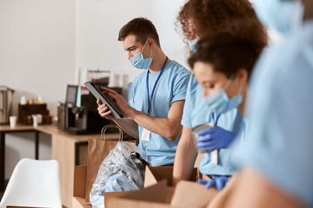 Group of busy volunteers wearing blue uniform, protective masks and gloves calculating, sorting...