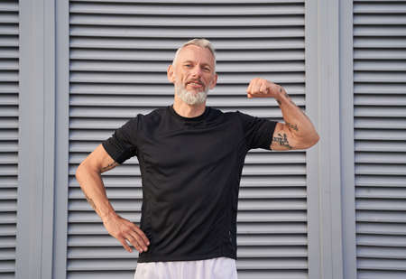 Confident middle aged man looking at camera, showing his bicep while posing outdoors over gray backgroundの写真素材