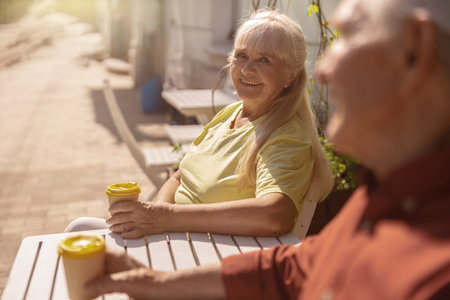 Happy blonde senior woman with paper cup and friend sit at small table in street cafeの写真素材