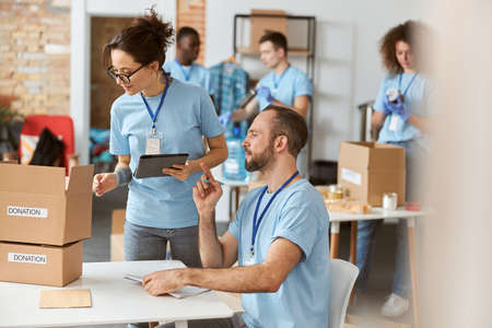 Female and male volunteers working together on donation project indoors. They are calculating, sorting and packing items in cardboard boxesの写真素材