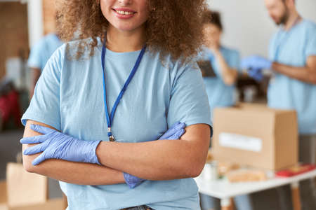 Cropped portrait of young woman, volunteer in blue uniform smiling at camera while standing with arms crossed indoors. Team sorting, packing items in the backgroundの写真素材