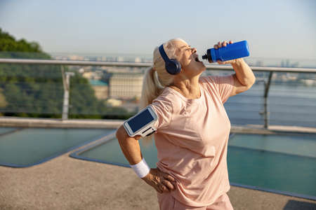 Blonde mature woman in pink tracksuit drinks water on footbridge in summerの写真素材
