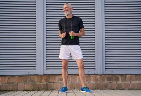 Cheerful middle aged athletic man in sportswear smiling aside while holding skipping rope, standing outdoors over grey backgroundの写真素材
