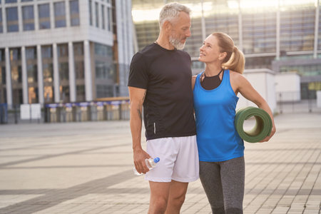 Beautiful mature couple, man and woman in sportswear walking together along the city street with yoga mat after morning workoutの写真素材