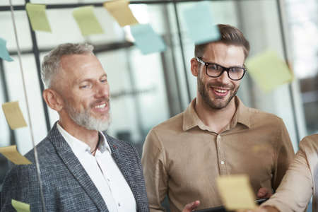 Two cheerful male coworkers standing in the modern office and looking at colorful sticky notes on a...