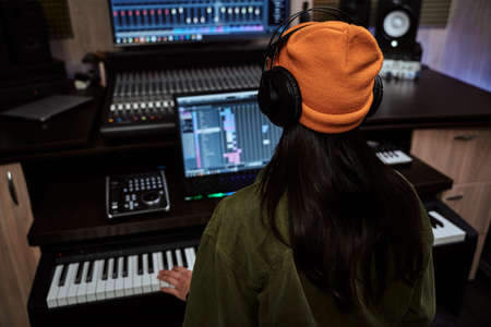 Young woman, female artist looking focused while playing keyboard synthesizer, creating music, sitting in recording studioの写真素材
