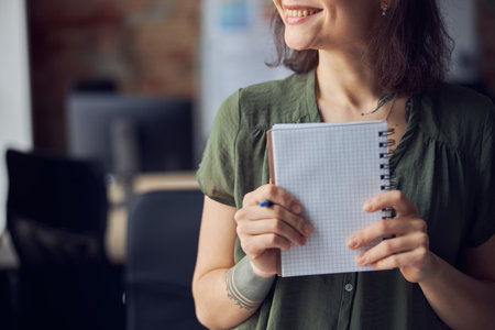 Cropped shot of young woman in casual wear holding empty notebook and pen in her hands and smiling aside. Interior designer or architect thinking about new project, standing in her officeの写真素材