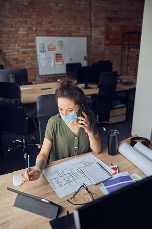 Busy interior designer wearing protective mask making a call while working with blueprint on new creative project, sitting at the table in her officeの写真素材