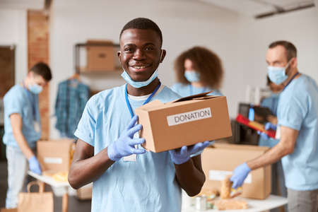 African american young male volunteer in blue uniform, protective mask and gloves smiling at camera while holding cardboard box for Donationの写真素材