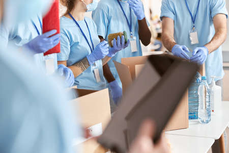 Cropped shot of volunteers in blue uniform, protective masks and gloves sorting, packing food and water in cardboard boxes, working together on donation projectの写真素材