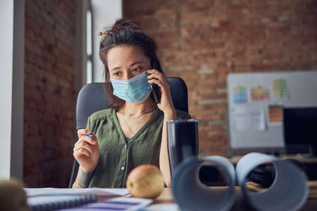 Portrait of serious interior designer or architect wearing protective mask talking on the phone while working on new poject, sitting at the table in her officeの写真素材