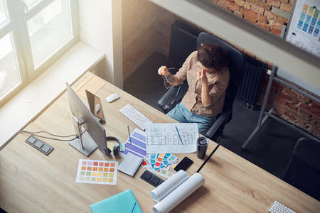 High angle view of tired interior designer woman taking off glasses and resting while working on new project, using pc in modern officeの写真素材