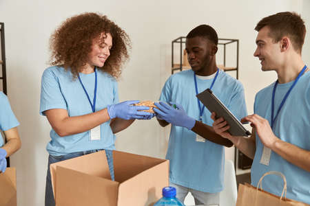 Team of diverse young volunteers in protective gloves smiling while sorting, packing foodstuff in cardboard box, working together on donation project indoorsの写真素材