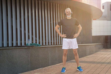 Full length shot of joyful sportive middle aged man in sportswear and headphones smiling while warming up, getting ready for workout outdoorsの写真素材
