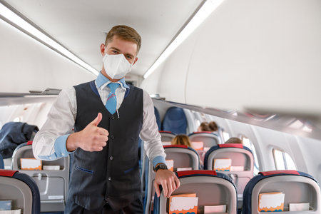 Male steward in medical mask doing thumbs up gesture in airplaneの写真素材