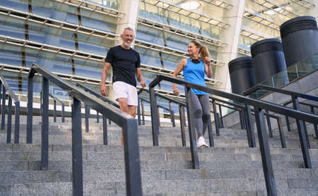 Joyful mature couple, man and woman in sportswear having workout together outdoors, standing on the stairs in cityの写真素材