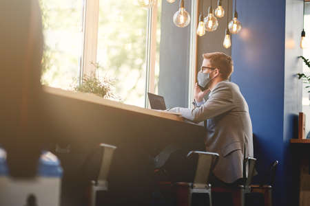 Young businessman wearing protective face mask talking by phone while working remotely in cafe during outbreakの写真素材