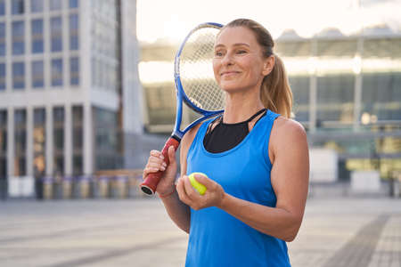 Beautiful middle aged woman, professional tennis player holding tennis racket and a ball ready for playing tennis in the cityの写真素材