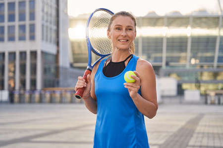 Portrait of attractive middle aged sportswoman, professional tennis player holding tennis racket and a ball ready for playing tennis in the cityの写真素材