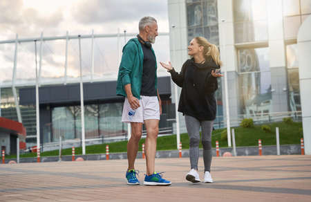 Full length shot of positive sportive middle aged couple in sportswear looking cheerful while talking, walking together outdoors after workoutの写真素材