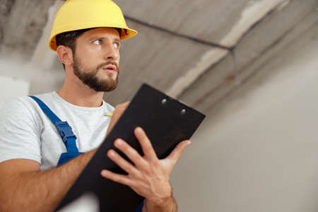 Pensive repairman in workwear and hardhat holding clipboard and writing down details of an order while standing indoors during renovation workの写真素材