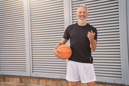 Joyful middle aged man in sportswear holding basketball ball and smiling at camera while standing outdoorsの写真素材