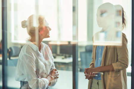 Two diverse female coworkers having informal conversation while standing in the modern officeの写真素材