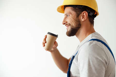 Side view portrait of smiling handyman in uniform and hard hat drinking coffee during a break while standing indoorsの写真素材