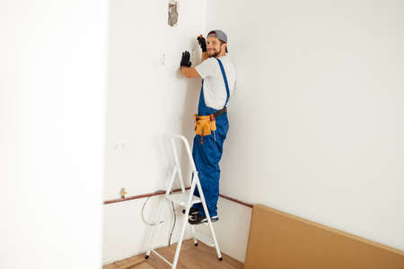 Enthusiastic electrician, handyman smiling at camera while working on installing socket on wall, standing on a ladderの写真素材