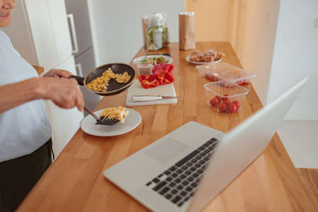 Pensioner female using laptop while cooking at kitchenの写真素材