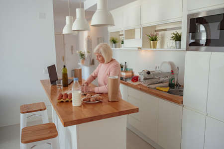 Diligent elderly woman making dough with flour and ingredients for baking while sitting at breakfast...