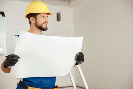 Portrait of thoughtful construction worker in hardhat looking away while studying blueprint, standing in new apartmentの写真素材