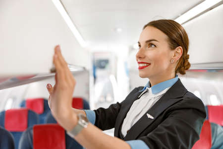 Smiling woman air hostess closing overhead luggage bin in aircraftの写真素材